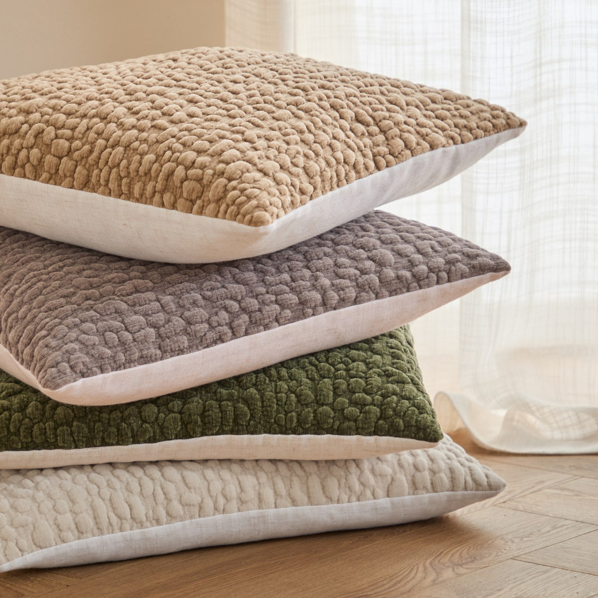 Stack of four Pebble Linen Chenille cushions with textured pebble-like chenille fronts and white linen backing, in beige, grey, olive green, and cream, shown on a wooden floor.