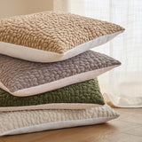 Stack of four Pebble Linen Chenille cushions with textured pebble-like chenille fronts and white linen backing, in beige, grey, olive green, and cream, shown on a wooden floor.
