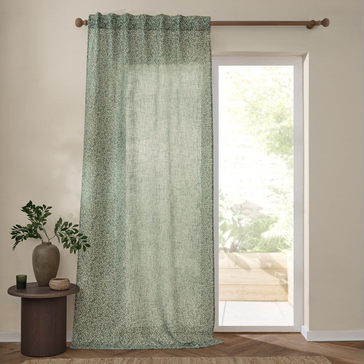 Perrie Leaf Floral Polyester Curtain Panel in forest green with a trailing white leaf print, hung from a wooden rod beside a glass door in a living room setting.