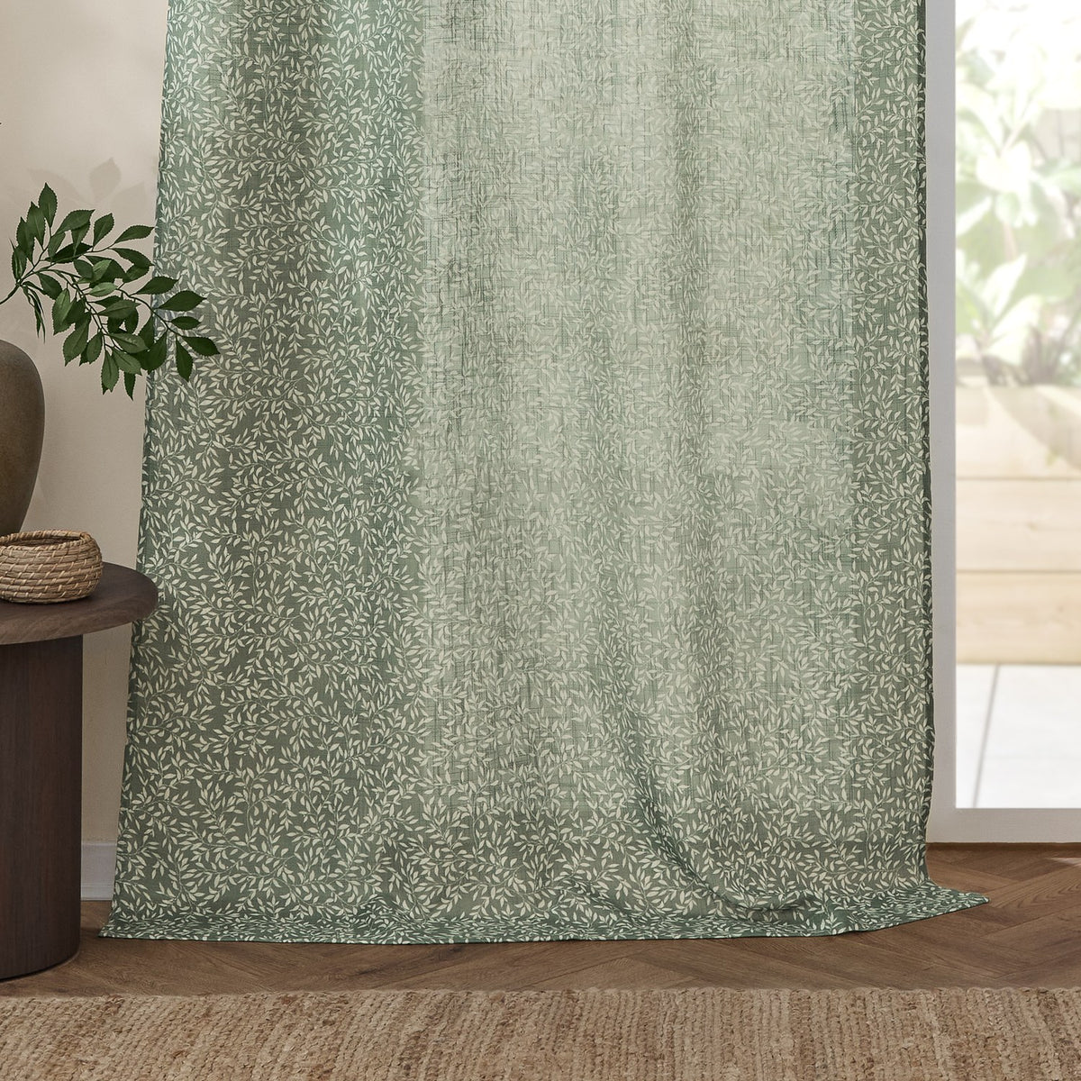 Perrie Leaf Floral Polyester Curtain Panel in Forest green, a sheer fabric panel with a trailing white leaf print, shown hanging beside a glass door in a living room setting.