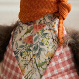 Close-up of the Phineas Polyester Door Stop, a sitting bird figure with a floral print jacket over pink gingham, a rust-orange knotted neck scarf, and a grey feathery tail.