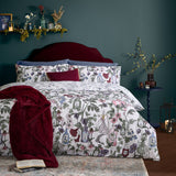 Pick Your Poison duvet cover set on a bed, off-white with floral apothecary bottle prints in pink, green, purple and red, styled with blue and burgundy cushions in a dark green bedroom.