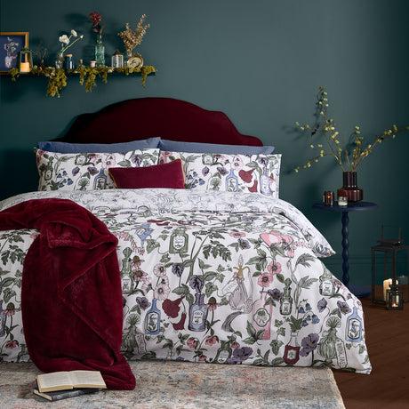 Pick Your Poison duvet cover set on a bed, off-white with floral apothecary bottle prints in pink, green, purple and red, styled with blue and burgundy cushions in a dark green bedroom.