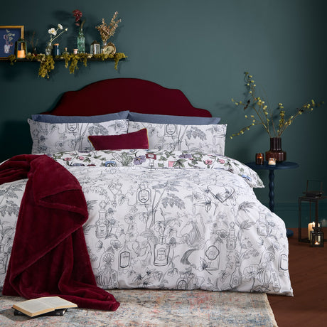 Pick Your Poison Duvet Cover Set on a bed with burgundy velvet headboard, showing an illustrated botanical and bottle print in multicolour with a red velvet cushion and throw in a dark green bedroom.
