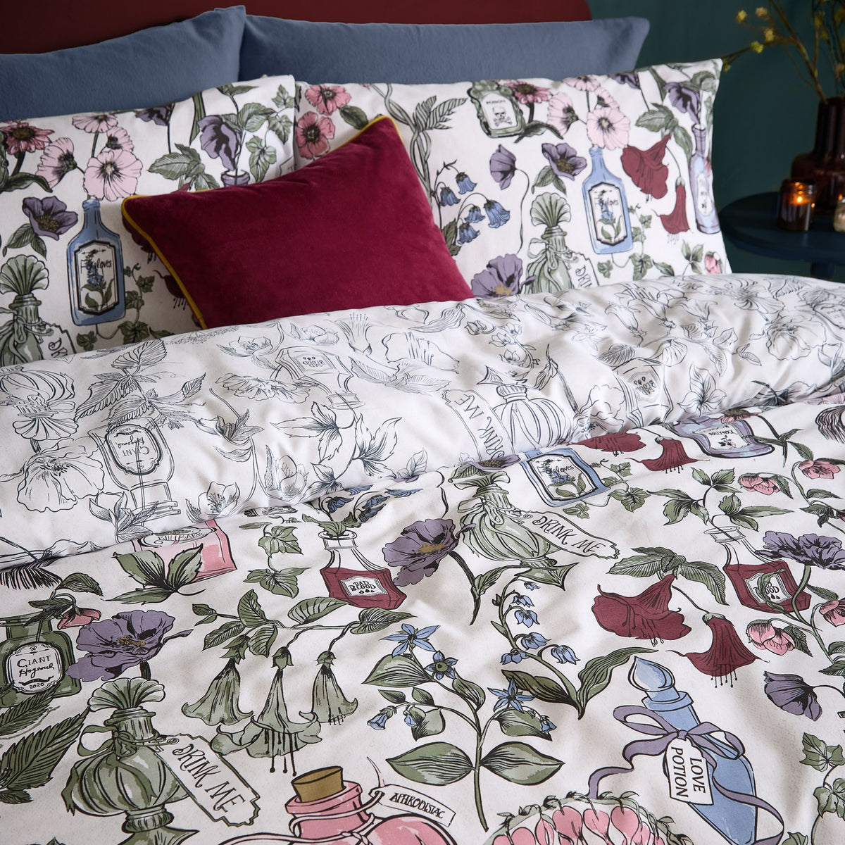 Pick Your Poison Duvet Cover Set on a bed, off-white ground with illustrated flowers and apothecary bottle labels in pink, green, purple and berry red, styled with a red velvet cushion.