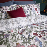 Pick Your Poison Duvet Cover Set on a bed, off-white ground with illustrated flowers and apothecary bottle labels in pink, green, purple and berry red, styled with a red velvet cushion.