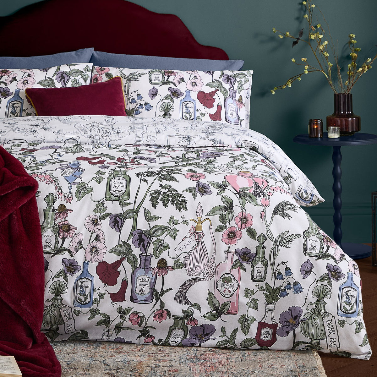 Off-white Pick Your Poison duvet cover set with pink, purple, green and red botanical florals and apothecary bottle print, styled with burgundy velvet cushions and a throw.