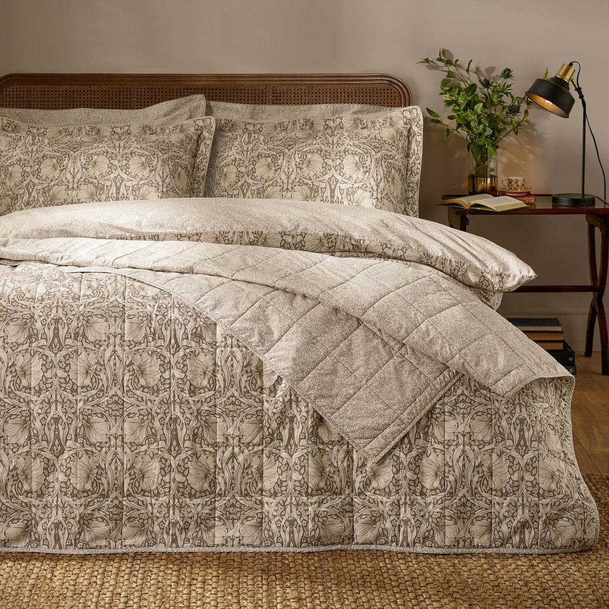 Pimpernel quilted cotton bedspread in natural and chocolate florals on a bed with matching pillowcases, styled in a bedroom with cane headboard and jute rug.