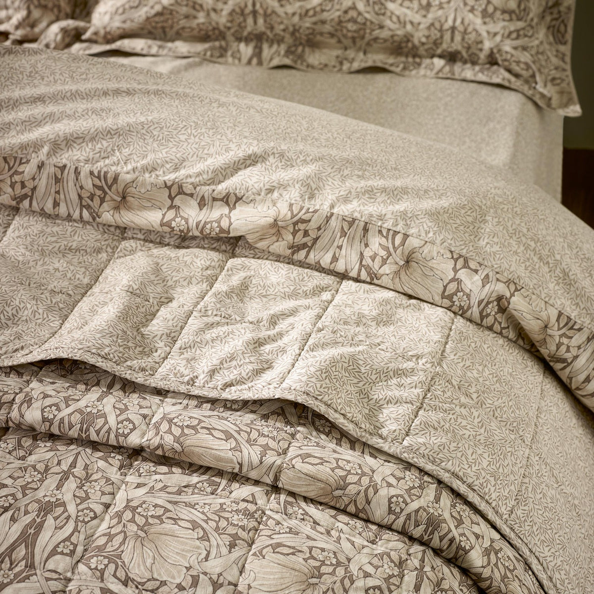 Pimpernel quilted cotton bedspread in natural and chocolate tones, shown folded on a bed with matching floral pillowcases, reverse leafy design visible at the turned edge.