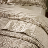 Pimpernel quilted cotton bedspread in natural and chocolate tones, shown folded on a bed with matching floral pillowcases, reverse leafy design visible at the turned edge.