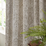 Pimpernel Jacquard Linen pencil pleat curtains in beige, showing the woven floral pattern in folds beside a window with a green plant in the foreground.