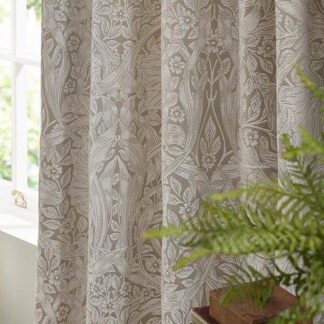 Pimpernel Jacquard Linen pencil pleat curtains in beige, showing the woven floral pattern in folds beside a window with a green plant in the foreground.