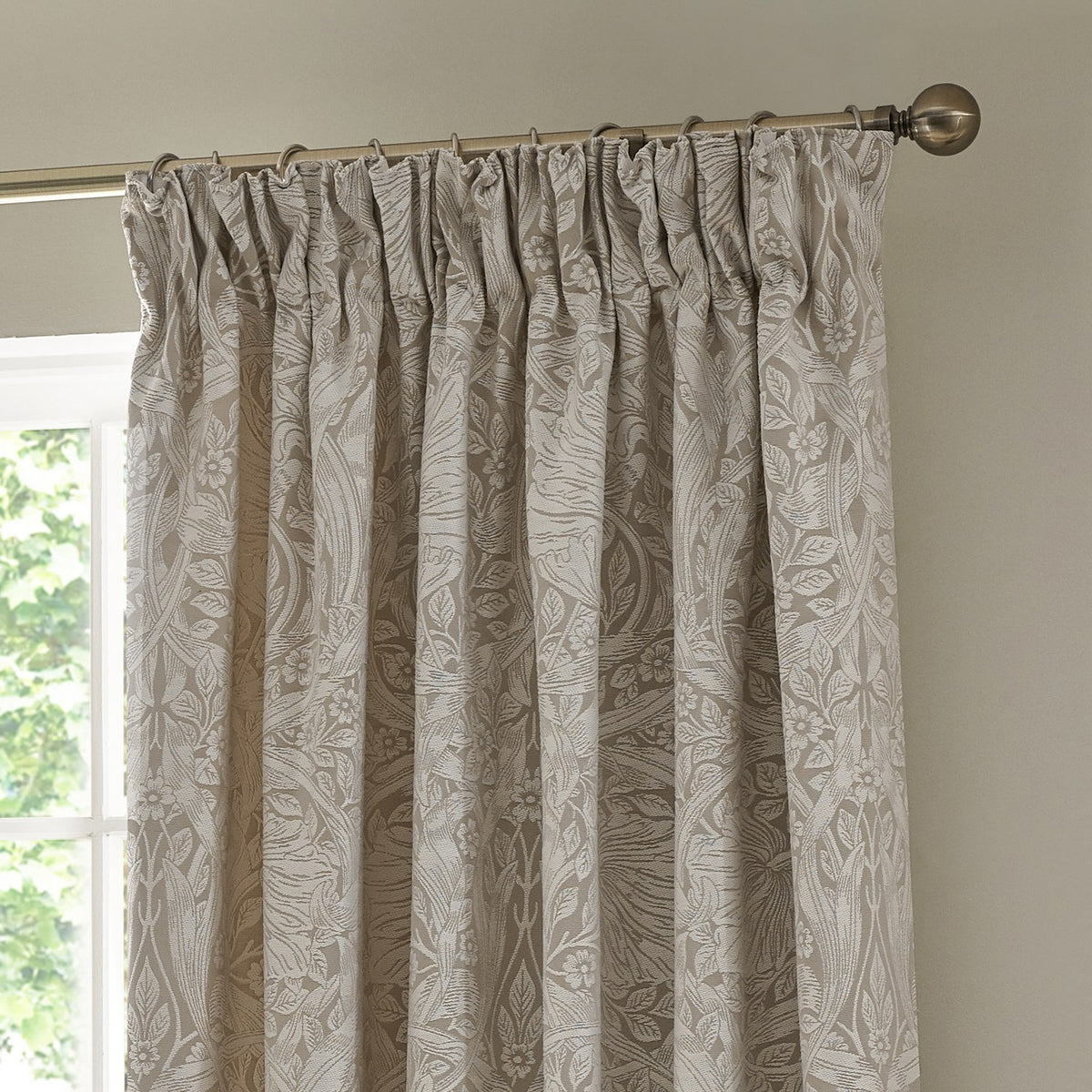 Pimpernel Jacquard pencil pleat curtains in linen beige, showing woven floral pattern and pleated header on rings, hung from a bronze-toned pole near a window.