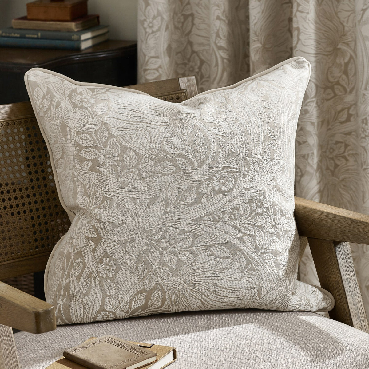 Pimpernel Jacquard Linen Cushion in cream with woven floral foliage pattern, piped edges, styled on a wooden armchair beside a curtain in matching print.