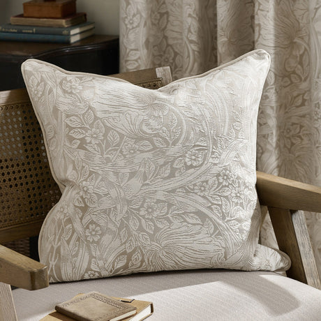 Pimpernel Jacquard Linen Cushion in cream with woven floral foliage pattern, piped edges, styled on a wooden armchair beside a curtain in matching print.