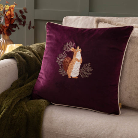 Pineberry Forest Animal Velvet Cushion in plum with embroidered red squirrel among foliage, cream piped edge, styled on a sofa with a green throw.