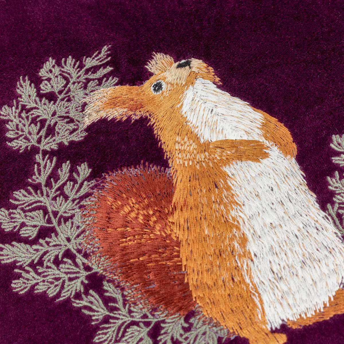 Close-up of the Pineberry Forest Animal Velvet Cushion in plum, showing a red squirrel embroidered amid grey-green foliage on a plum velvet ground.