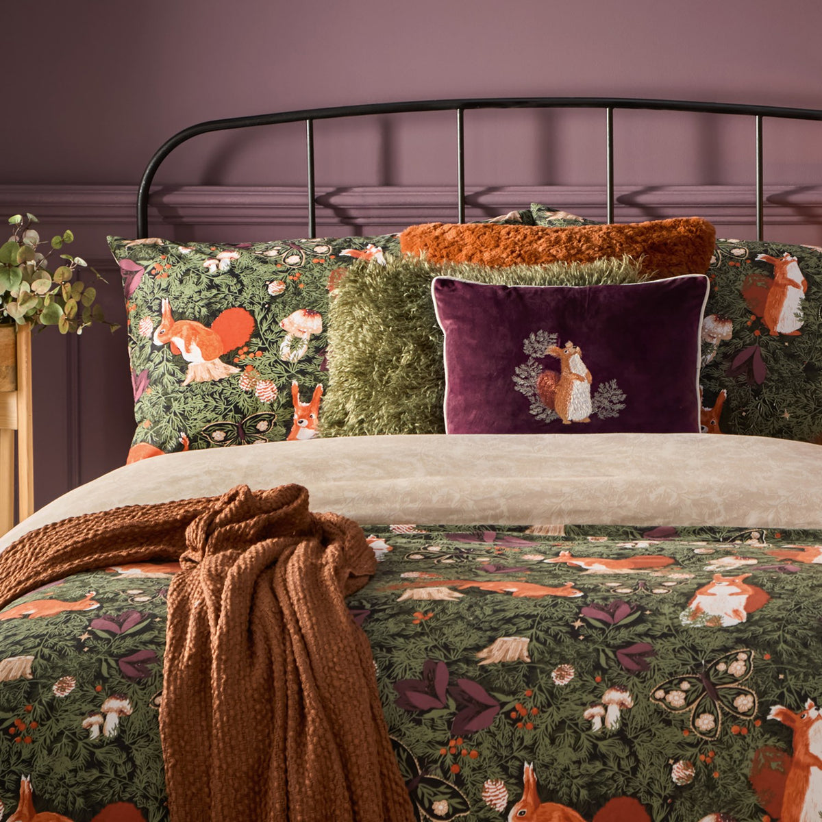 Plum velvet rectangular cushion with embroidered red squirrel and foliage design, styled on a forest-print bed with rust throw, green shaggy cushion and orange textured bolster in a bedroom setting.