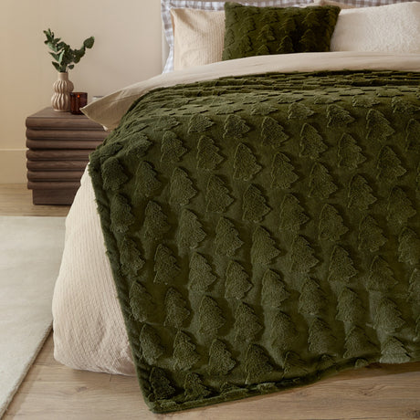 Pines Velvet Faux Fur Throw in forest green draped over a bed, showing raised tree-textured pile alongside cream bedding and a matching cushion.