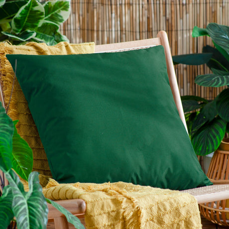 Plain Printed Polyester Cushion in bottle green, square 55 x 55cm design, shown on a wooden chair with a mustard throw and greenery in an outdoor setting.