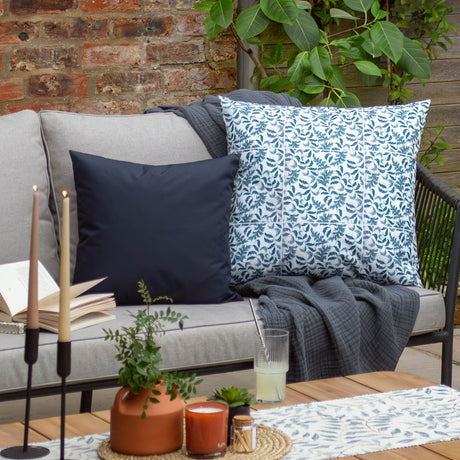 Navy Plain Printed Polyester Cushion on a grey outdoor sofa, styled with a blue leaf-patterned cushion and dark throw against a brick wall.