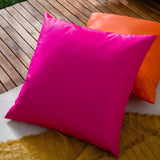 Plain Printed Polyester Cushion in bright pink with a satin-like sheen, square shape, shown outdoors on a wooden deck beside an orange cushion.