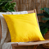 Plain Printed Polyester Cushion in bright yellow, 55 x 55cm square shape, shown on a rattan chair in an outdoor garden setting with plants and a woven fence behind.