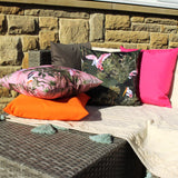 Platalea Jungle Polyester Cushion in bottle green with pink spoonbill and stork print, styled outdoors among pink, orange and pink cushions on a bench.