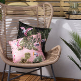 Platalea Jungle Polyester Cushion in Blush pink with spoonbill and stork print, shown on a woven rattan chair alongside a matching dark green cushion.