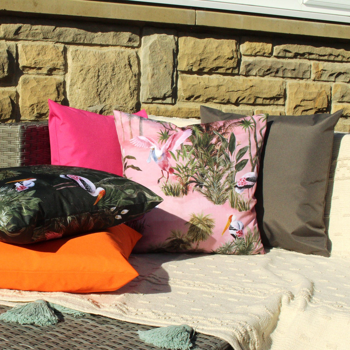 Platalea Jungle Polyester Cushion in Blush, a square pink cushion printed with wading spoonbills and tropical foliage, shown outdoors with pink, orange, black and grey scatter cushions.