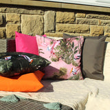 Platalea Jungle Polyester Cushion in Blush, a square pink cushion printed with wading spoonbills and tropical foliage, shown outdoors with pink, orange, black and grey scatter cushions.