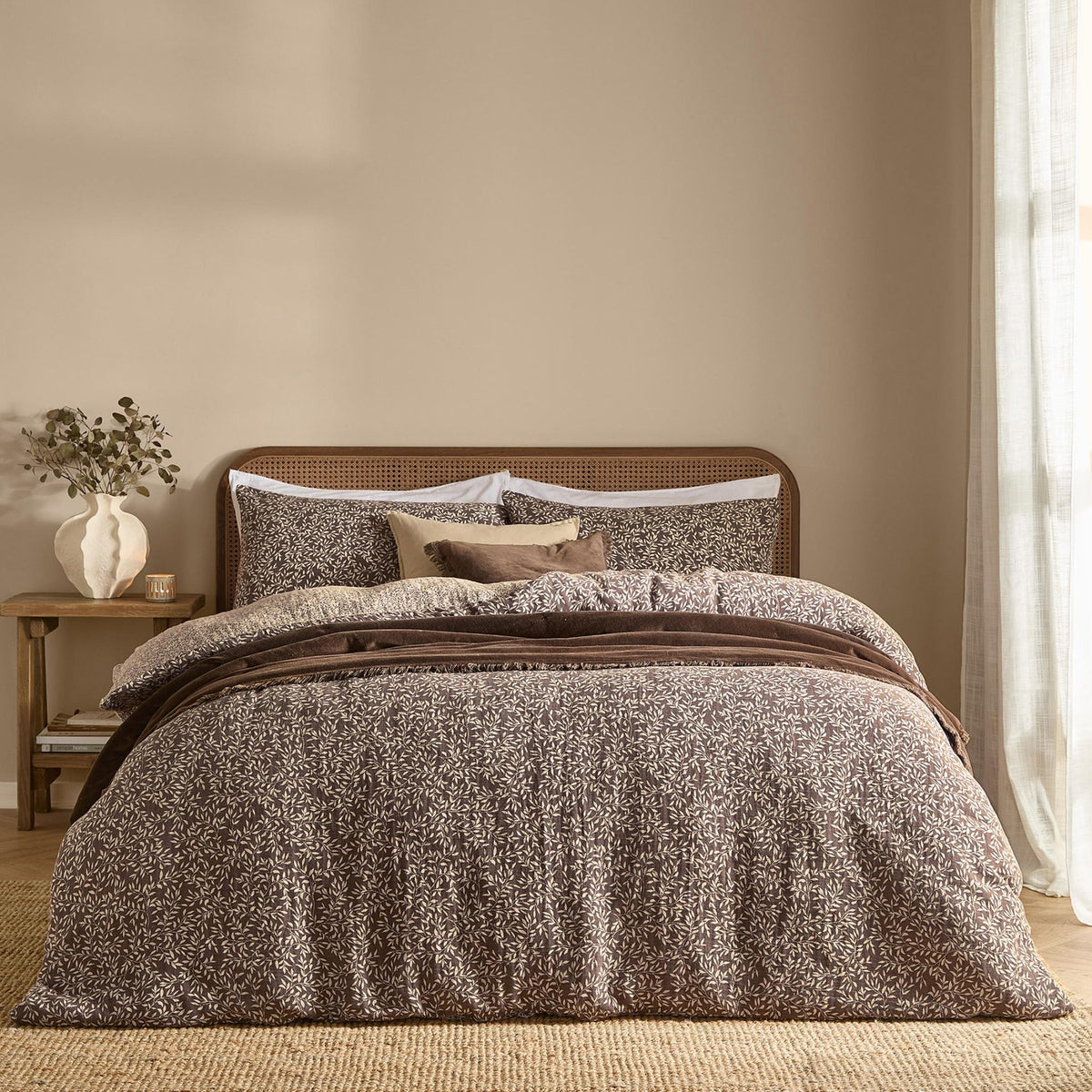 Perrie Leaf Cotton Ditsy Duvet Cover Set in Chocolate dressing a bed with matching pillowcases, cream leaf print on brown cotton, styled with cushions in a beige bedroom.