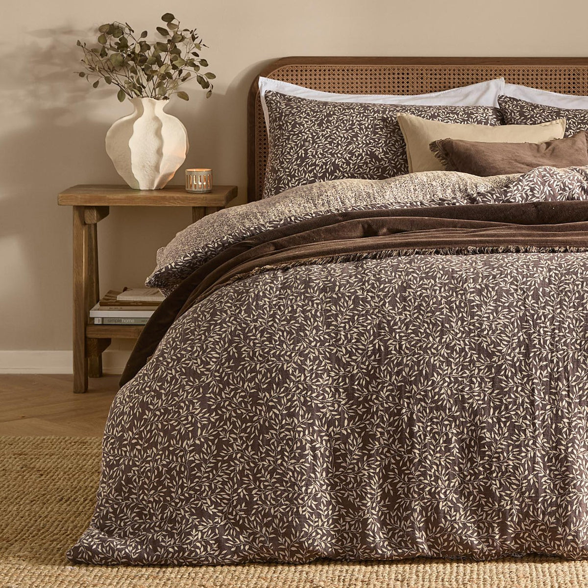 Perrie Leaf Cotton Ditsy Duvet Cover Set in chocolate brown with cream leaf print, styled on a bed with matching pillowcases, cushions and a cane headboard in a bedroom setting.