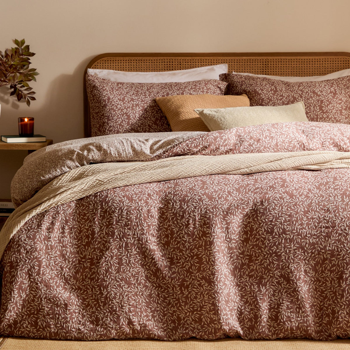 Perrie Leaf Cotton Ditsy Duvet Cover Set in cinnamon on a bed with matching pillowcases, cream ditsy leaf print, cane headboard, and layered cushions in a warm-toned bedroom.