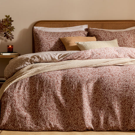 Perrie Leaf Cotton Ditsy Duvet Cover Set in cinnamon on a bed with matching pillowcases, cream ditsy leaf print, cane headboard, and layered cushions in a warm-toned bedroom.