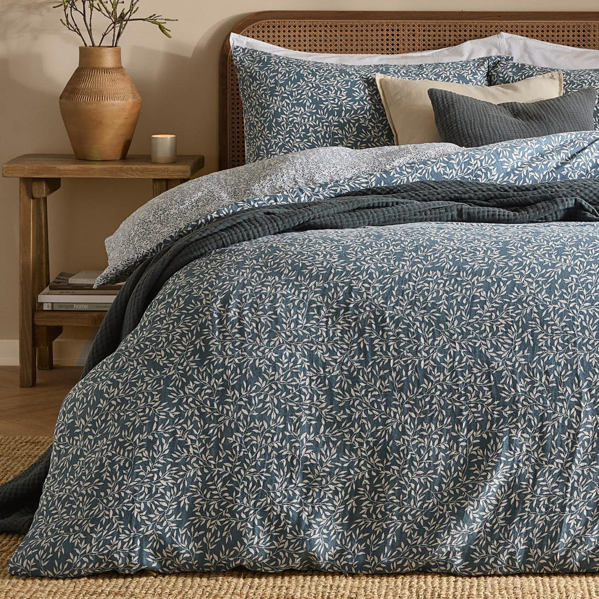 Perrie Leaf Cotton Ditsy Duvet Cover Set in denim blue with cream trailing leaf print, shown on a bed with matching pillowcases and dark grey quilted cushions in a bedroom setting.