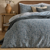 Perrie Leaf Cotton Ditsy Duvet Cover Set in denim blue with cream trailing leaf print, shown on a bed with matching pillowcases and dark grey quilted cushions in a bedroom setting.