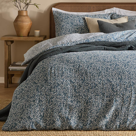 Perrie Leaf Cotton Ditsy Duvet Cover Set in denim blue with cream trailing leaf print, shown on a bed with matching pillowcases and dark grey quilted cushions in a bedroom setting.