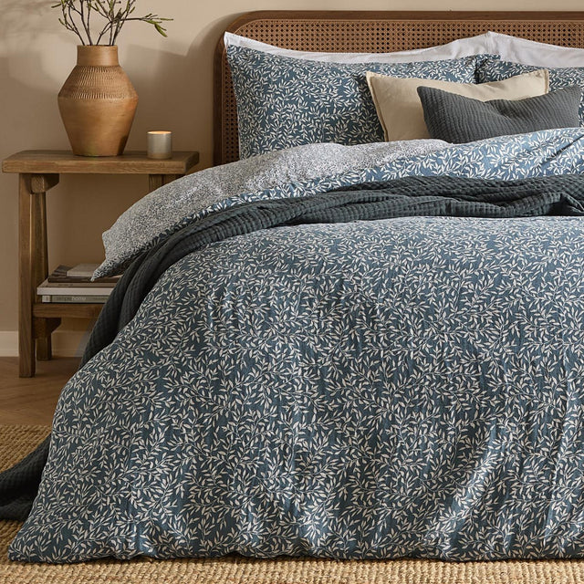 Perrie Leaf Cotton Ditsy Duvet Cover Set in denim blue with cream trailing leaf print, shown on a bed with matching pillowcases and dark grey quilted cushions in a bedroom setting.