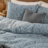 Perrie Leaf Cotton Ditsy Duvet Cover Set in denim blue with cream leaf-and-stem print, shown on a bed with matching pillowcases and a grey textured cushion.