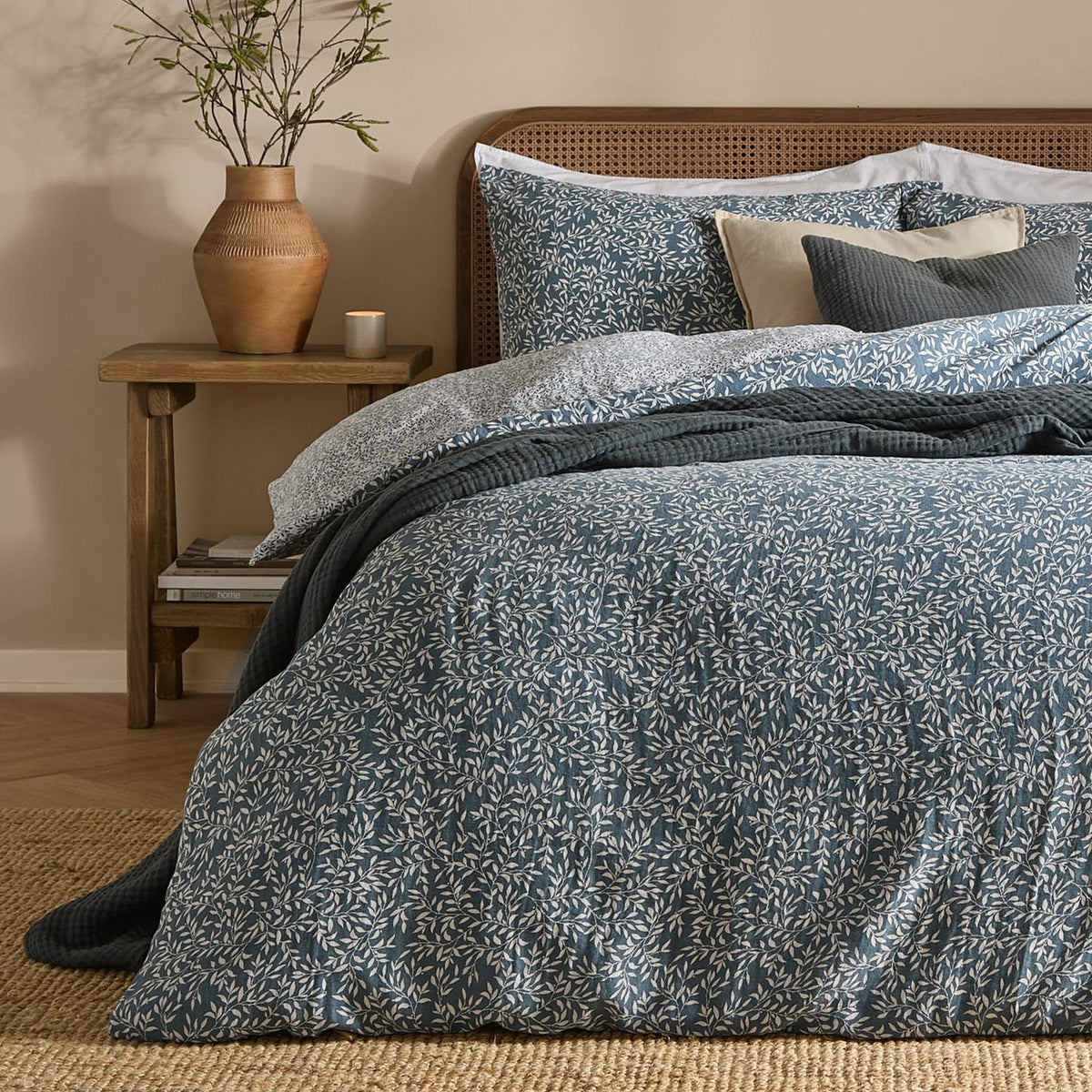 Perrie Leaf Cotton Ditsy Duvet Cover Set in denim blue with cream trailing leaf print, styled on a bed with cane headboard, patterned pillowcases and dark knitted throw in a bedroom setting.
