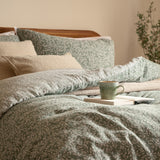 Perrie Leaf Cotton Ditsy Duvet Cover Set in Forest, styled on a bed with cream textured pillows, ditsy leaf print in green on cotton, alongside a mug and book.