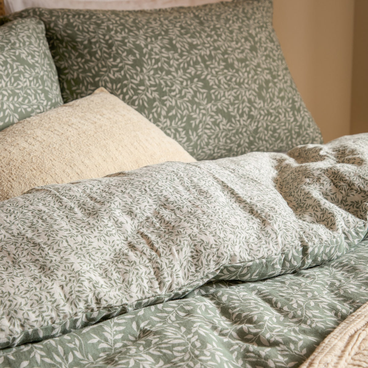 Perrie Leaf Cotton Ditsy Duvet Cover Set in Forest, cotton bedding with a green leaf print on a natural ground, shown with matching pillowcases and a plain cream cushion on a bed.