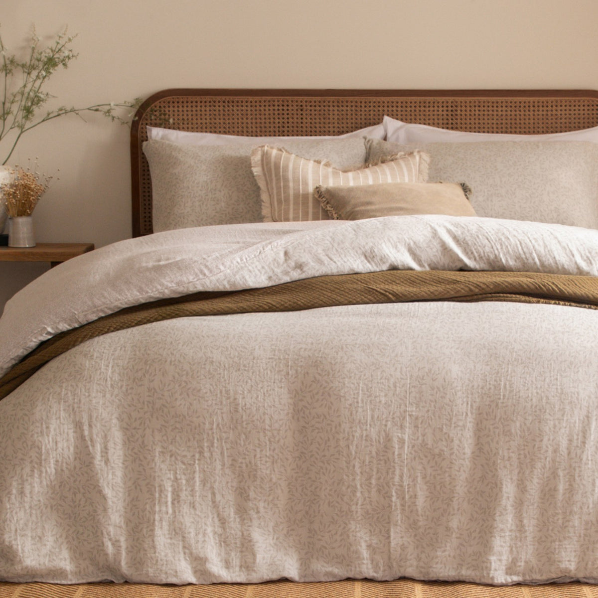 Perrie Leaf Cotton Ditsy Duvet Cover Set in Natural, styled on a bed with a rattan headboard, layered pillows, and a mustard textured throw in a bedroom setting.