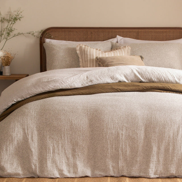 Perrie Leaf Cotton Ditsy Duvet Cover Set in Natural, styled on a bed with a rattan headboard, layered pillows, and a mustard textured throw in a bedroom setting.