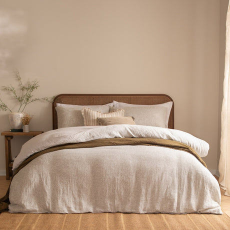 Perrie Leaf Cotton Ditsy Duvet Cover Set in Natural, styled on a bedroom bed with cane headboard, layered cushions, and an olive throw over the pale leaf-print cotton bedding.