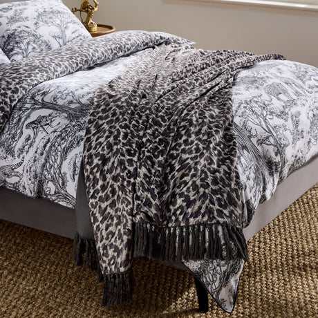Pokot Animal Velvet Throw in Monochrome, leopard-print grey and black velvet with knotted fringe edges, draped over a patterned bed in a bedroom setting.
