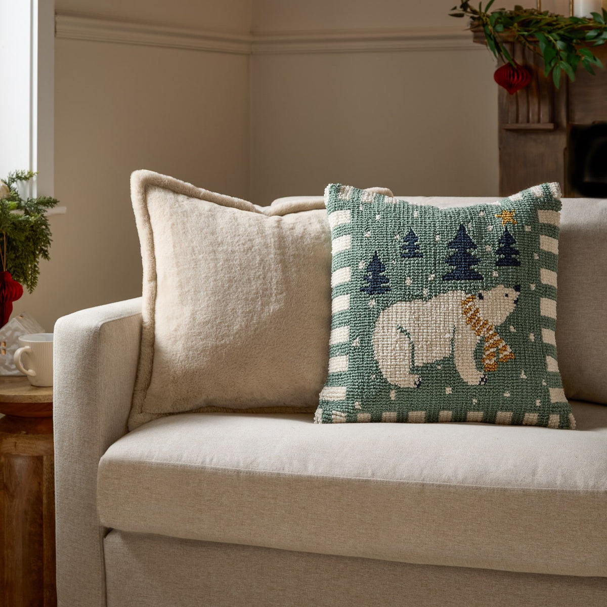 Square 45cm loop-tufted cushion in frosty blue-green with a polar bear, navy trees and gold star design, striped border, shown on a sofa beside a cream textured cushion.