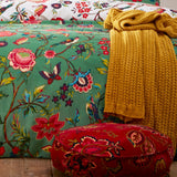 Pomelo Printed Floral Duvet Cover Set in Verdi green on a bed, with a mustard knit throw and red floral round cushion styled alongside on a wooden bench.