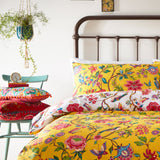 Pomelo Printed Floral Duvet Cover Set in yellow, polycotton bedding with red, pink, blue and green blooms, birds and butterflies, styled on a bed with a dark metal headboard.